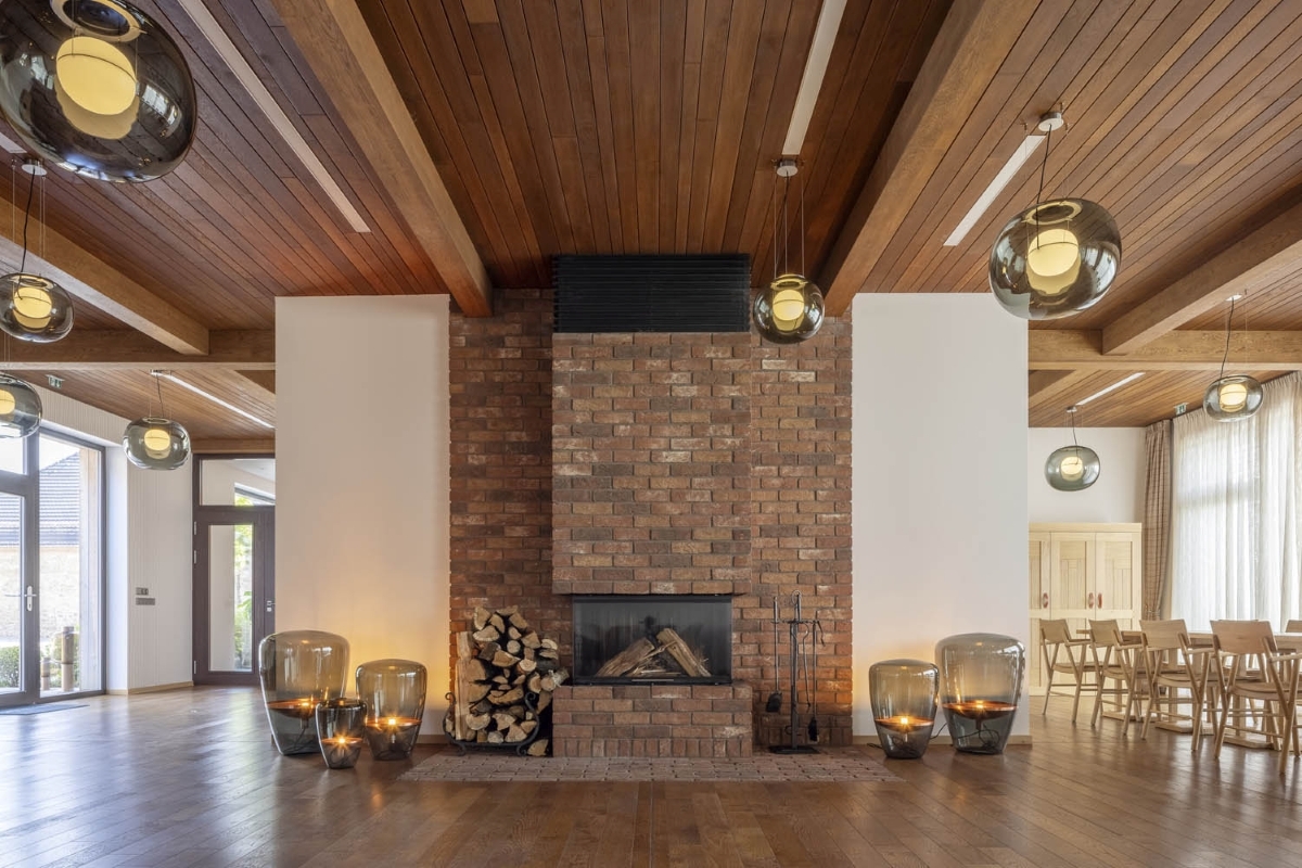 A living room with exposed beams, a central brick fireplace, Brokis Big One pendants and Balloons, and expansive windows.