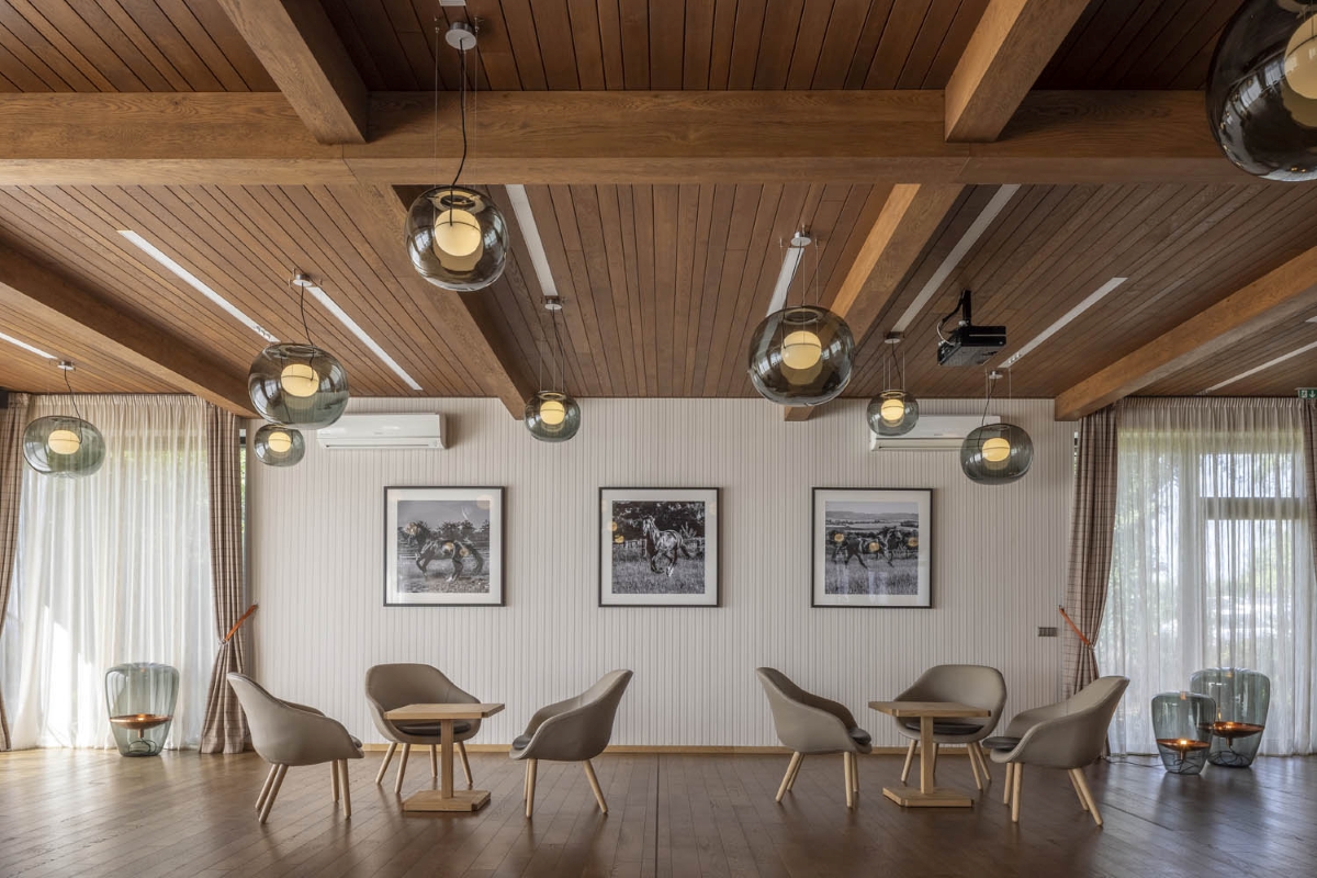 A lounge with wooden floors, Brokis Big One Large pendants, four beige chairs, sheer curtains, and striking equine art.