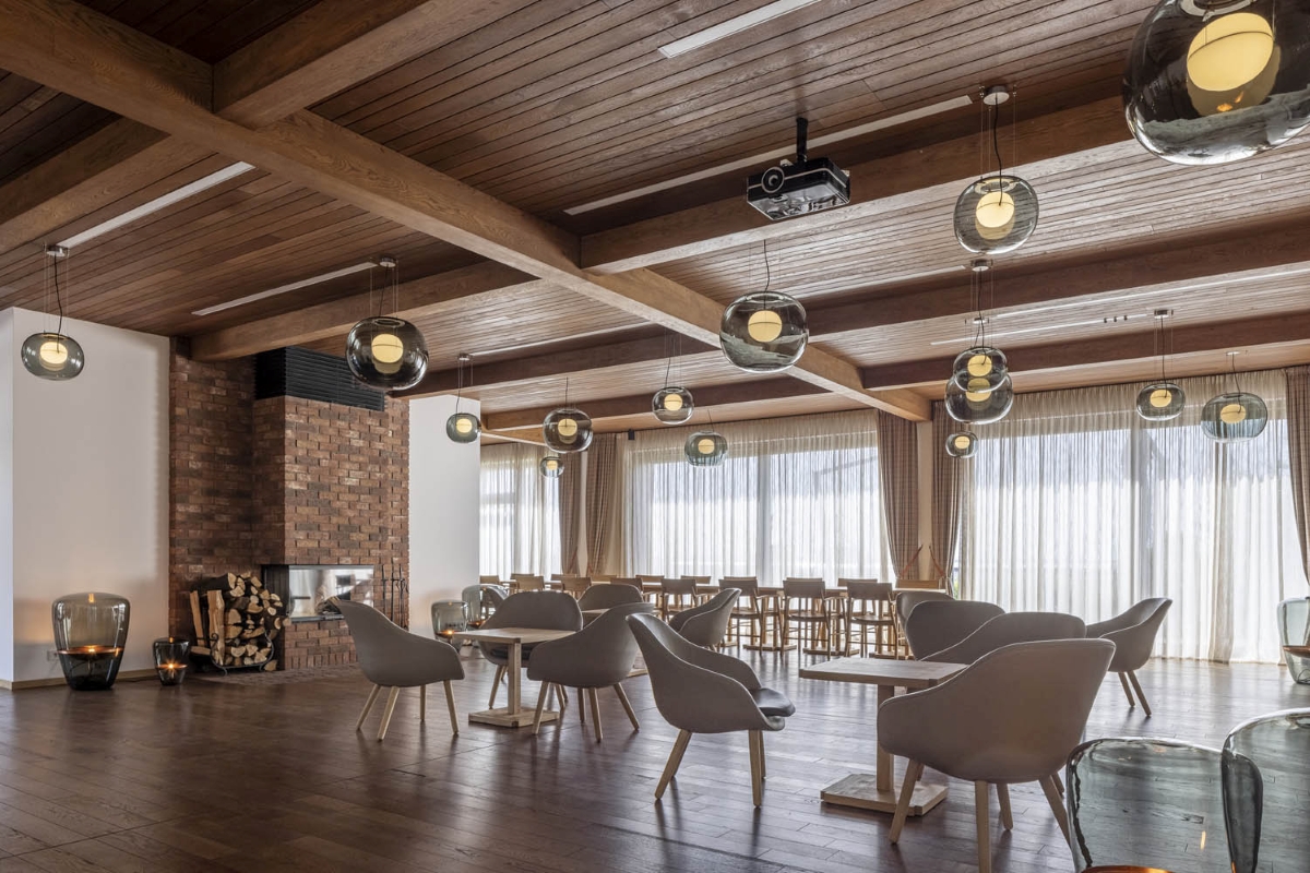 Lounge with wooden ceiling and floor, Brokis Big One Large pendants, round tables, grey chairs, fireplace, sheer-lit windows.