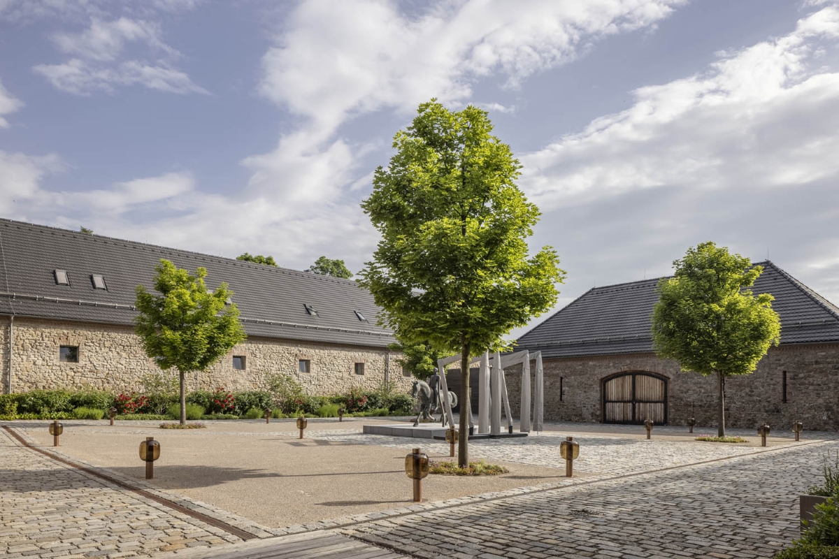A cobblestone courtyard with Brokis Bonbori Outdoor and a horse sculpture, framed by stone buildings and arched wooden doors.
