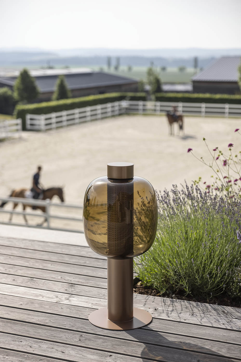A copper Bonbori Outdoor Large on a wooden decking framed by lavender, as riders glide past white-fenced paddocks at dusk.