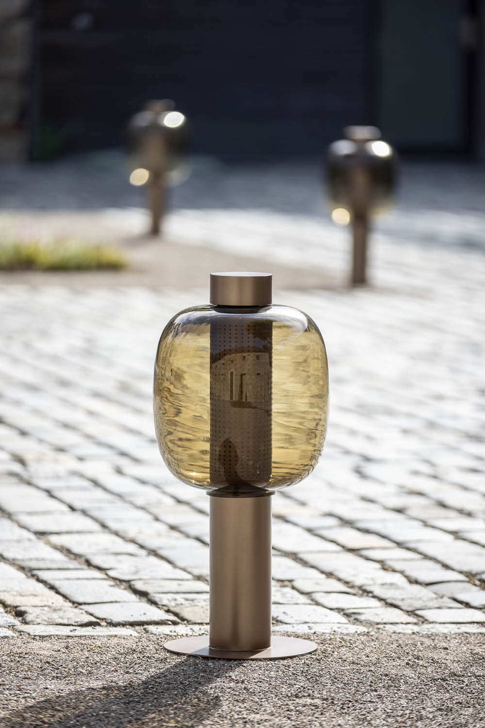 Brokis Bonbori Outdoor Large in smoke brown glass and copper stands on cobbles, others elegantly aglow behind.