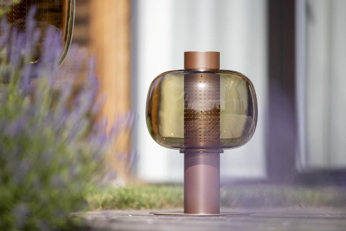 A Brokis Bonbori Outdoor Small with a copper base and brown glass shade on stone, framed by blurred purple flowers.