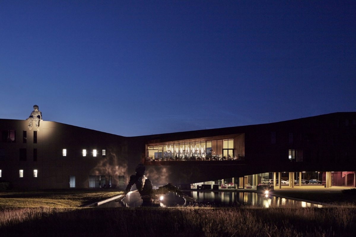 Hotel Miura at dusk glows with expansive windows. An astronaut sculpture perches above a misty sculpture and reflecting pond.