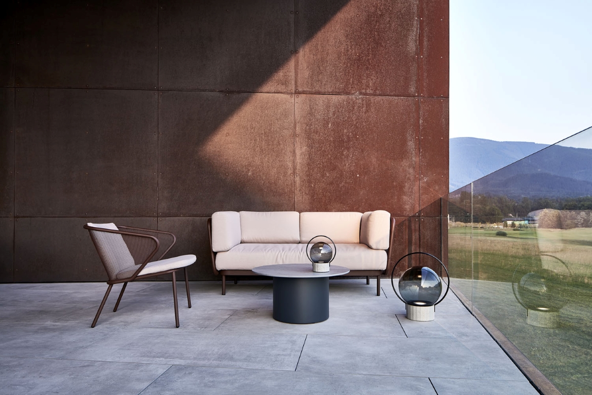 A terrace with a beige sofa, mesh chair, sculptural Brokis Sfera Portable lamps, dark table, and sweeping mountain views.