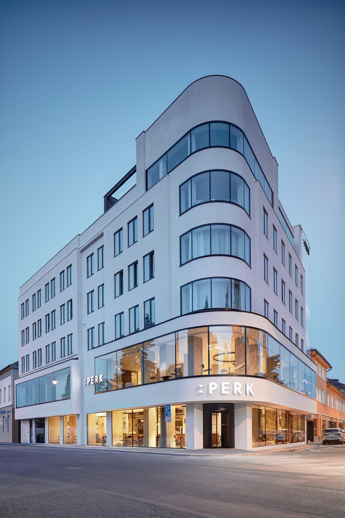 Hotel Perk sleek white multi-storey building with curved glass corners; interior glows warmly at dusk.