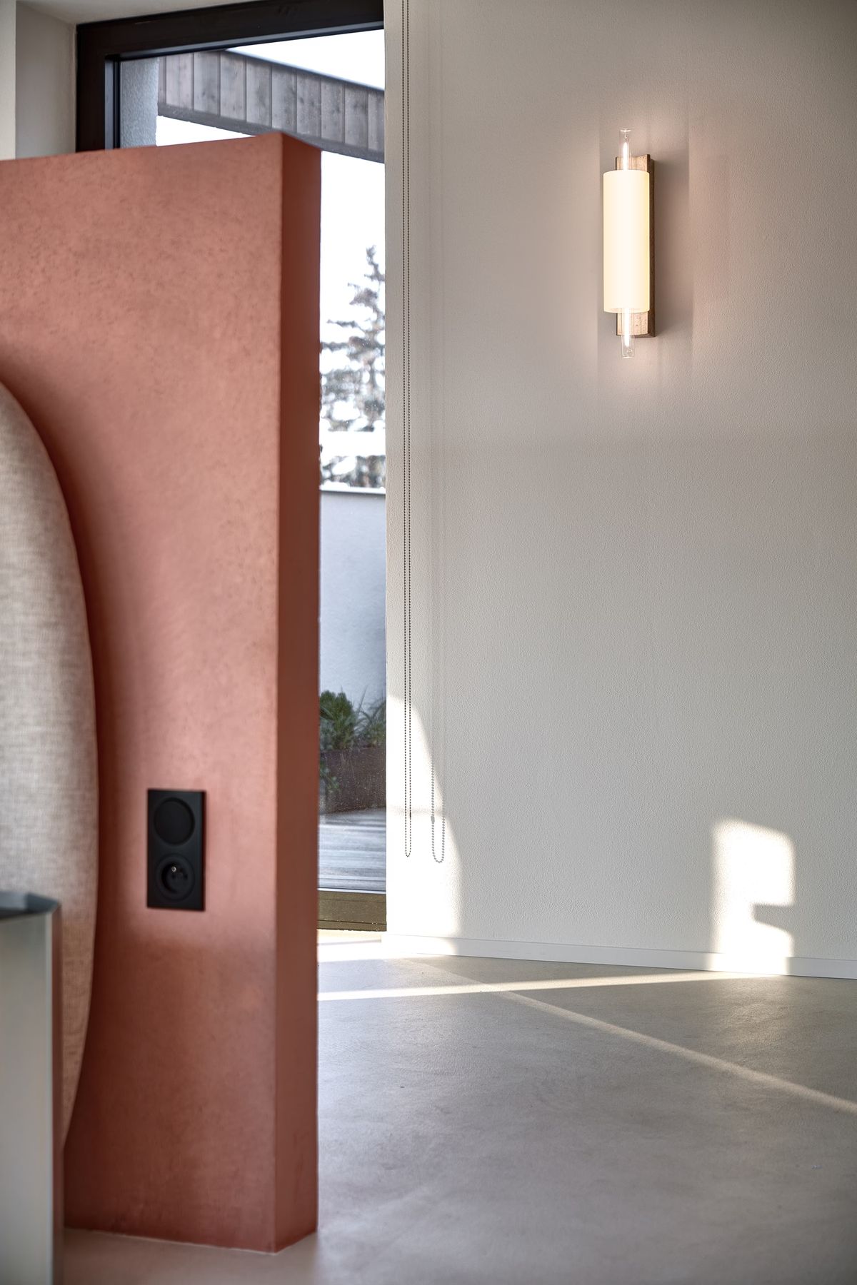 A modern, minimal interior with a blush pink partition and a Brokis Overlay Wall M light. Light fills the airy space.