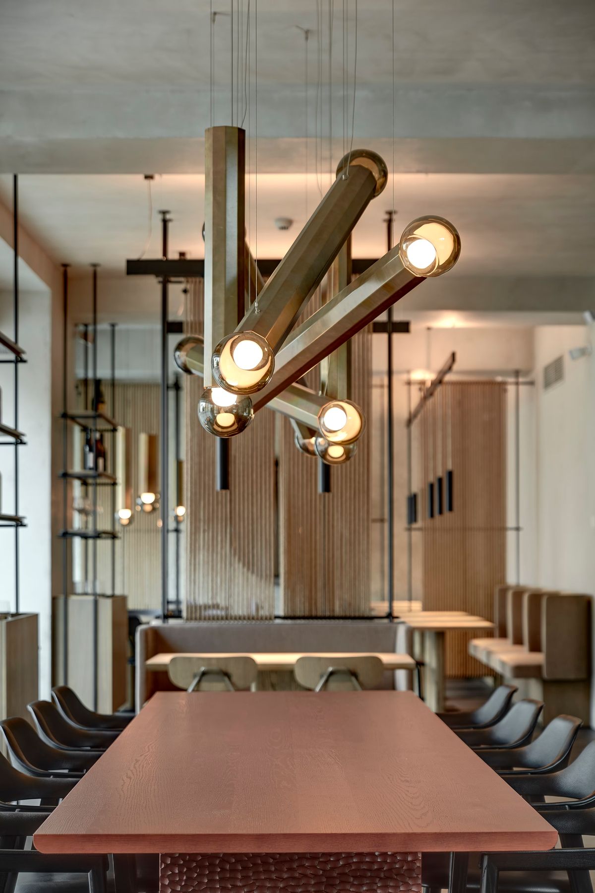 A modern dining area with a long wooden table, black chairs, and Brokis Prisma chandeliers with exposed glass bulbs.