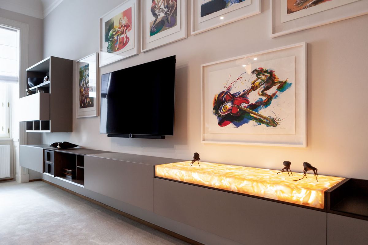 A luxurious living room with wall-mounted TV, sleek grey cabinetry, abstract art, and a BROKISGLASS backlit surface.