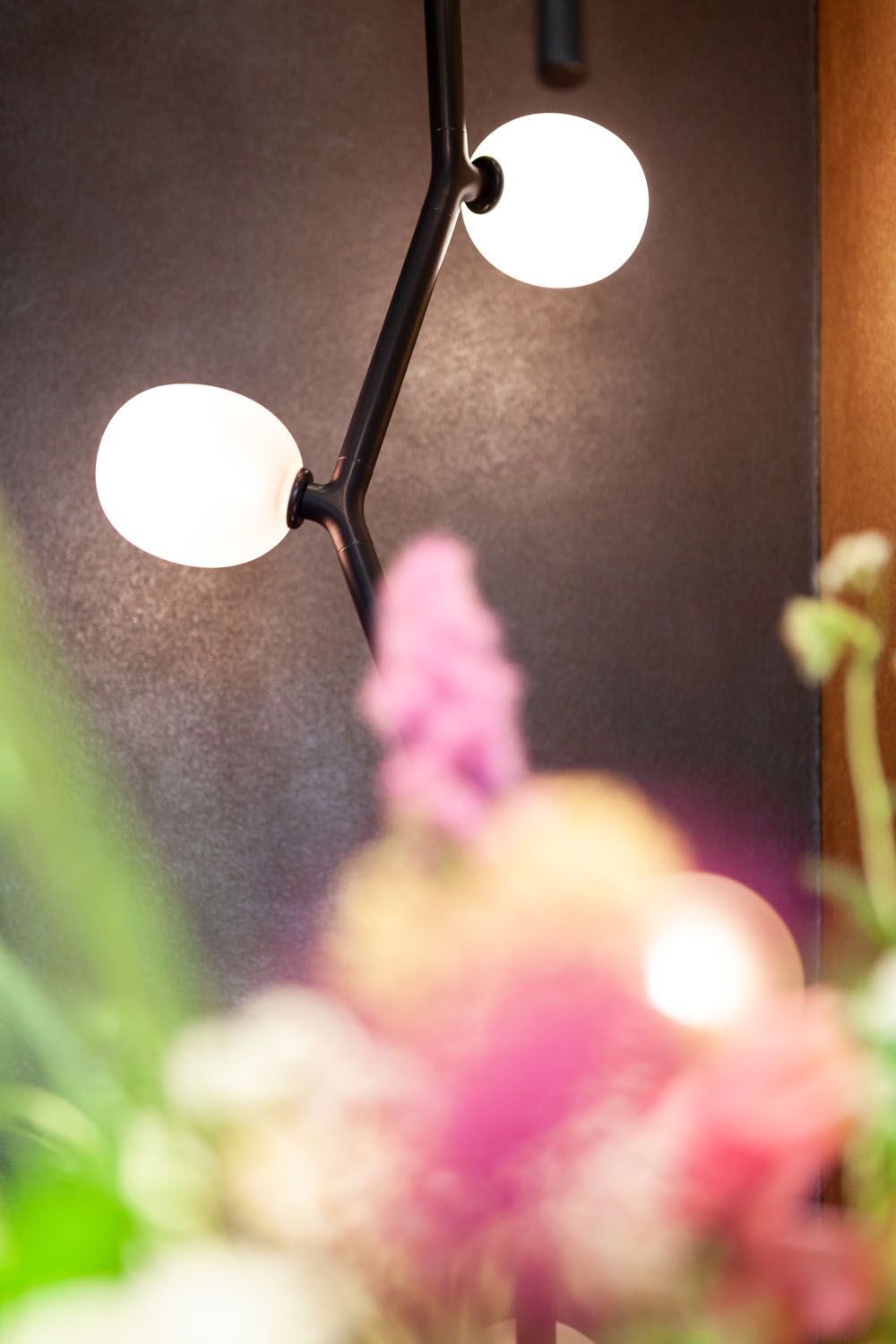 A Brokis Ivy wall light with two round white glass orbs glows against a dark wall, blurred florals evoking luxe warmth.