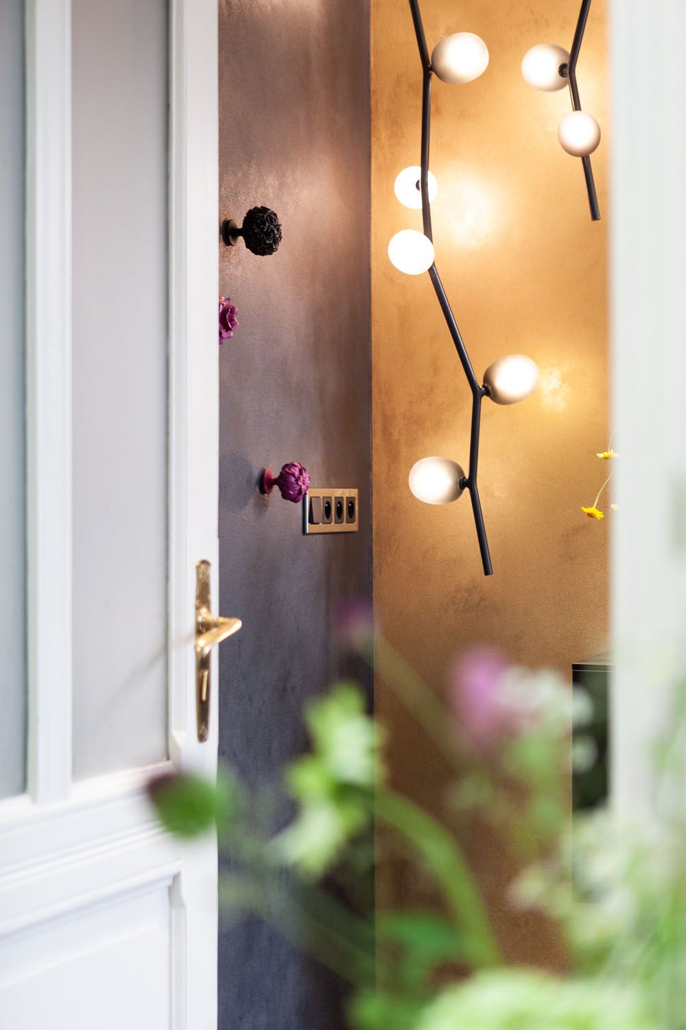 A white door opens to Brokis Ivy Vertical Pendants and artful purple wall hooks; greenery blurs in front.