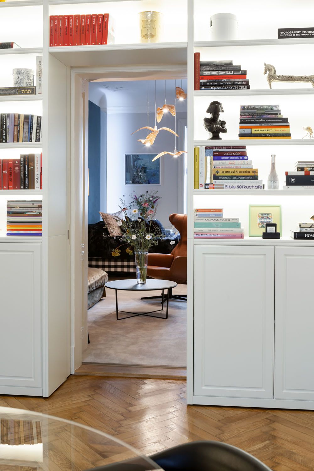 Bookcase walls open to a lounge with luxurious seating, Brokis Night Birds, stacked books, and herringbone wooden floors.
