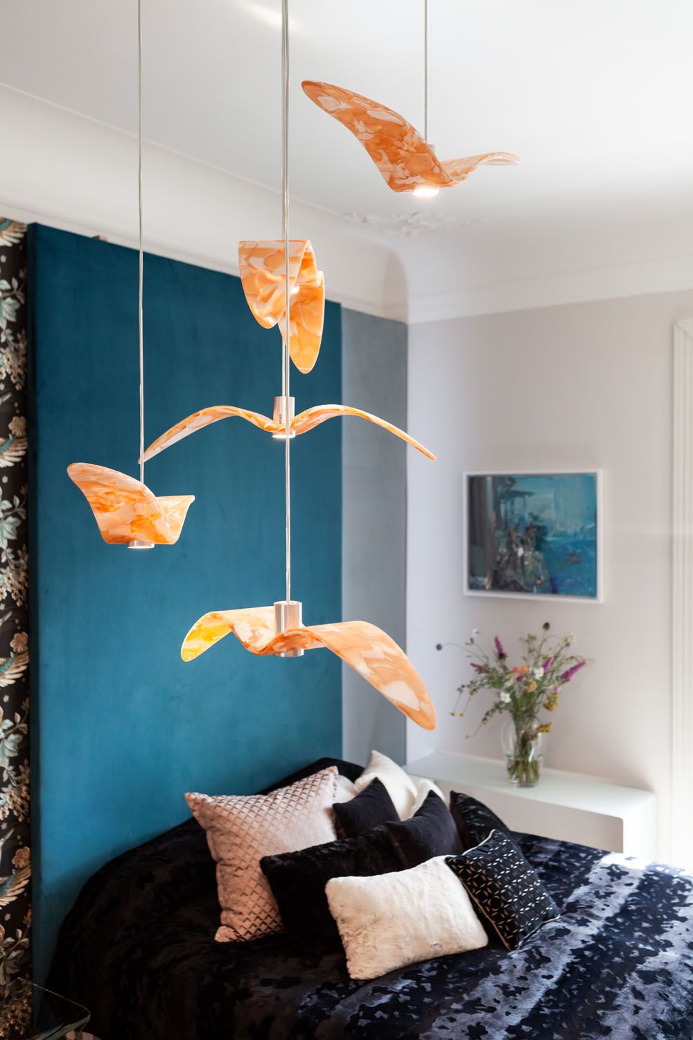 Brokis Night Birds pendants soar above a luxurious bed; blue wall, florals, and art add elegance.