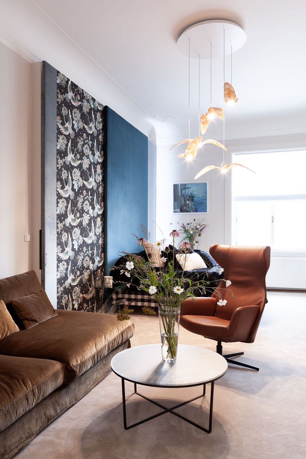 Modern living room with brown velvet sofa, leather armchair, Brokis Night Birds pendants, and large window.