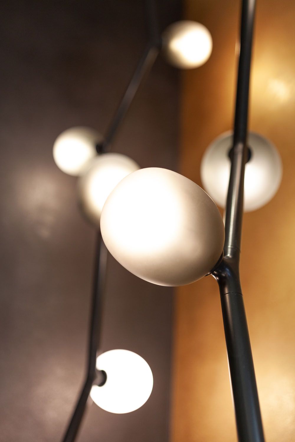 Close-up of a Brokis Ivy Vertical with handblown glass globes on sleek rods, glowing against a blurred golden backdrop.