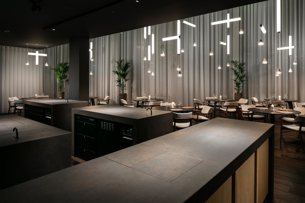 A restaurant interior with dim light, Brokis Puro pendants and cross-shaped lights, cream chairs, and lush potted plants.