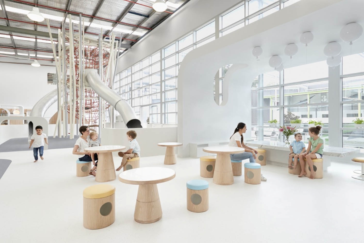 Children and an adult gather at round tables in a luminous play area with Brokis Memory ceiling lights overhead.