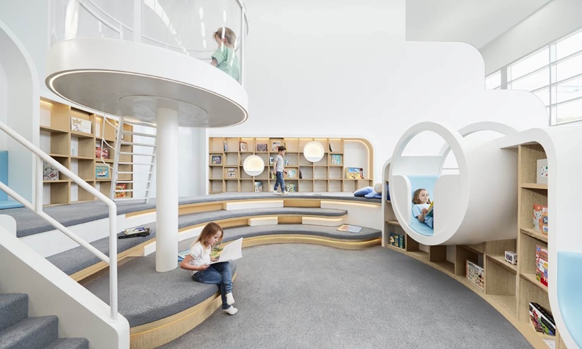 A luminous children’s library with curved shelves, cosy nooks, a circular pod, and tiered seating.