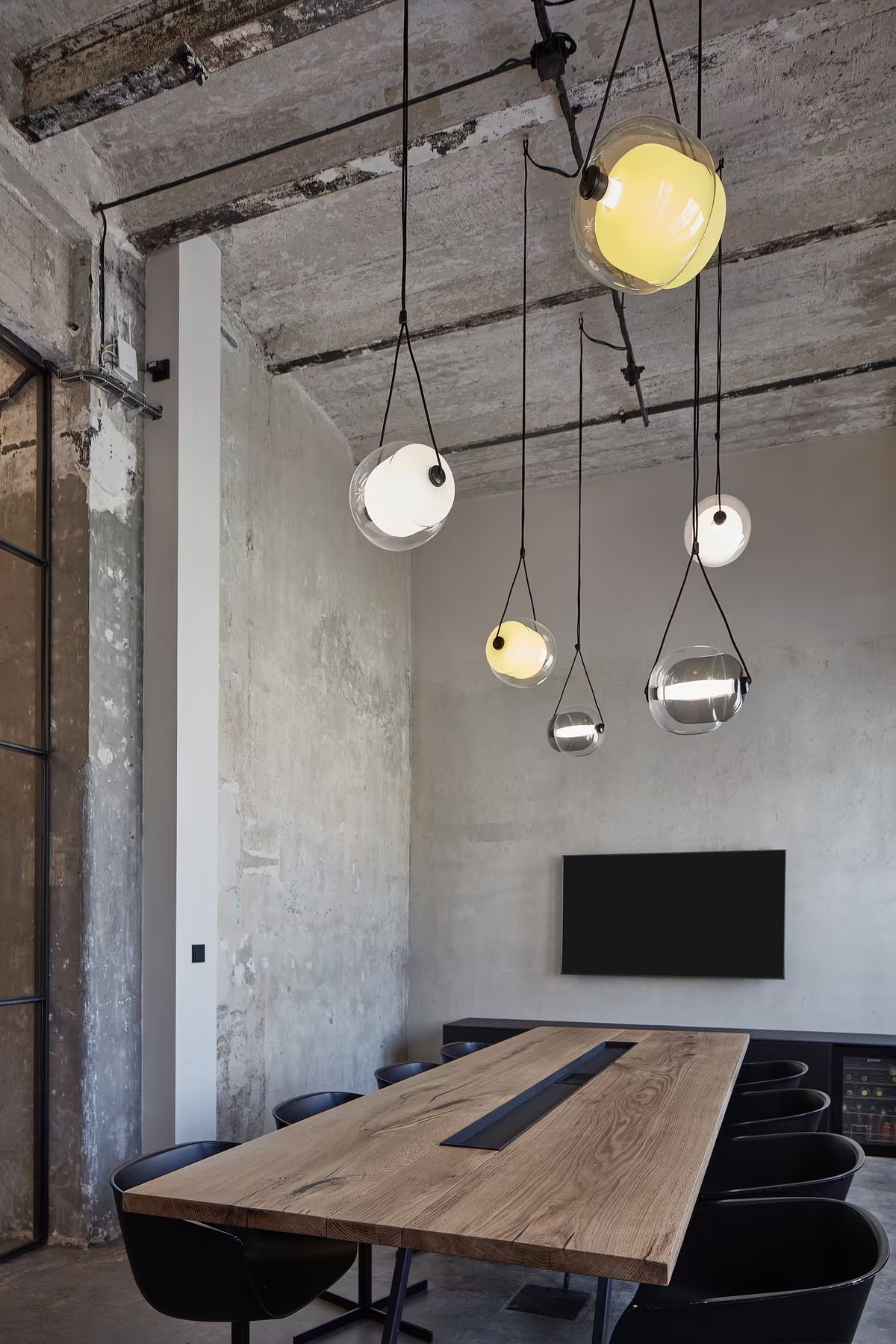 A modern meeting room with a long wooden table, black chairs, exposed concrete and Brokis Capsula Single pendants.