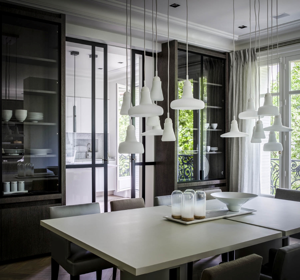 A sleek dining room with a white table, grey chairs, Brokis Shadows pendants, glass cupboards, and expansive windows.