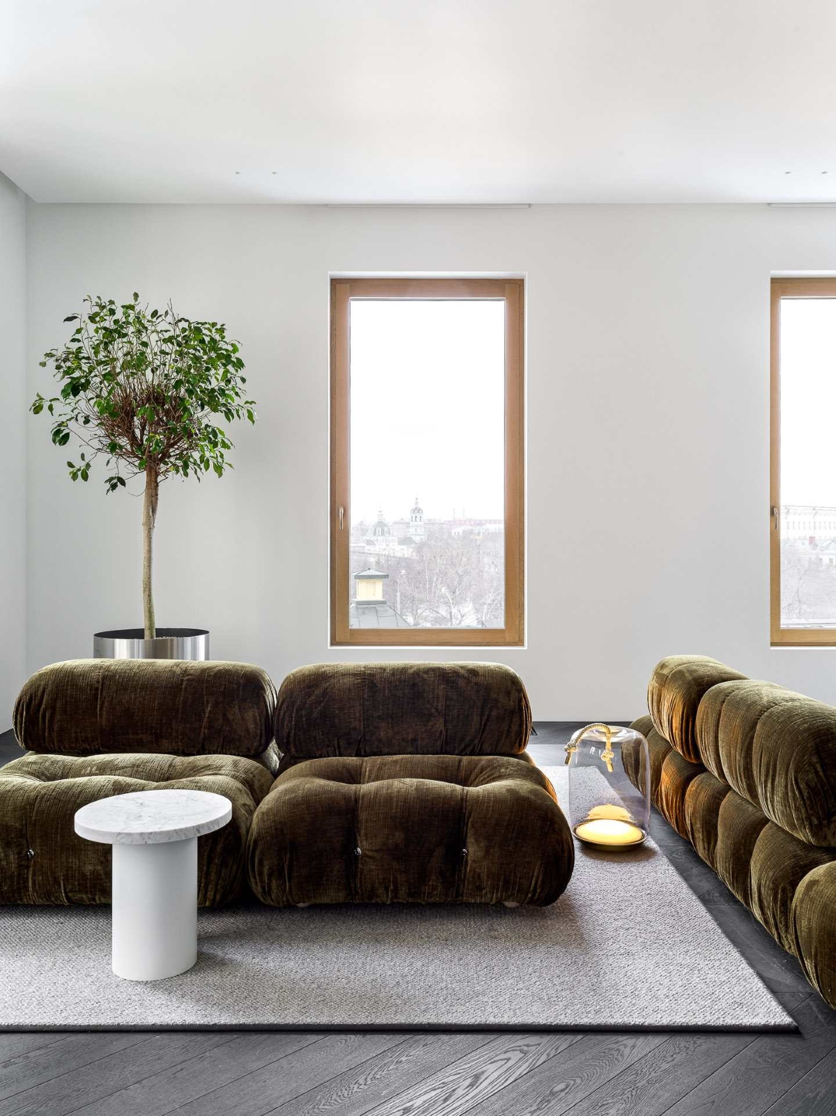 Chic living room with olive tufted sofas, grey rug, Brokis Knot Cilindro Table Battery, and potted tree by city-view windows.