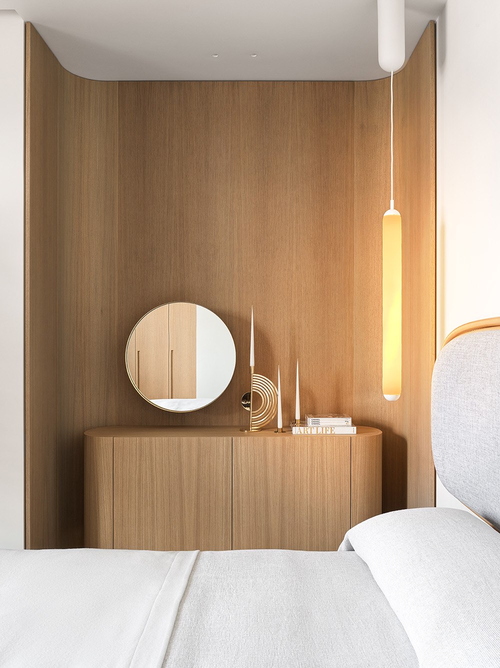 A minimalist bedroom with a light wood cupboard, curved panelling, round mirror, and Brokis Puro pendant over grey bedding.