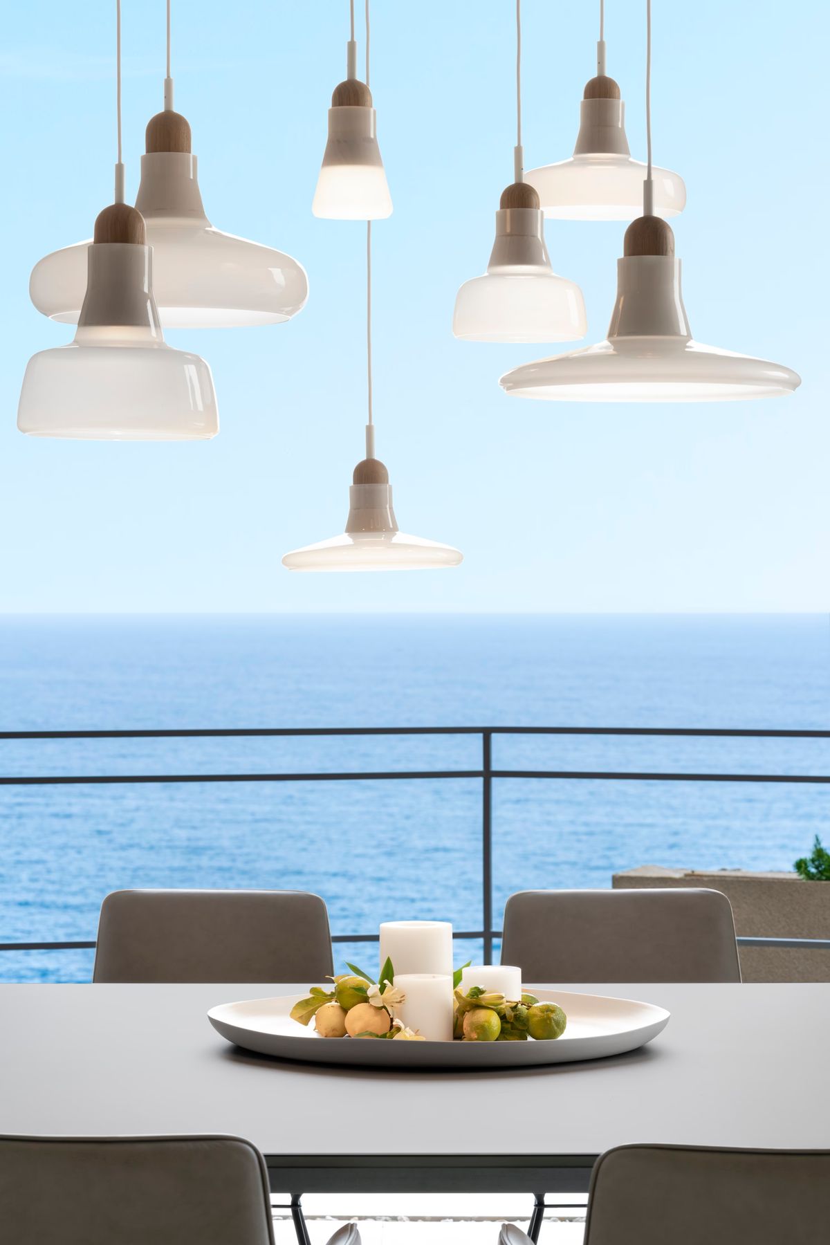 A modern dining area with grey chairs, a white tray centrepiece, and white Brokis Shadows pendants over a sea-view balcony.