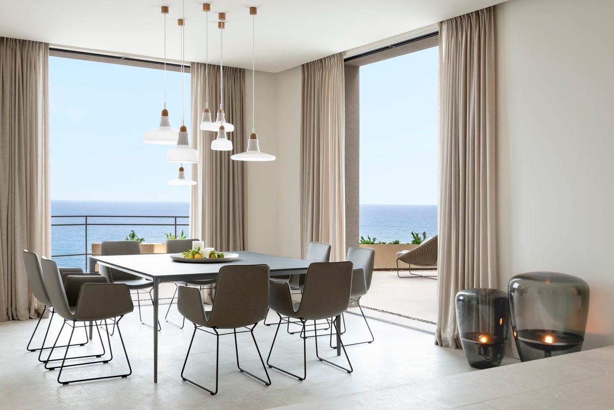 Modern dining room with sea view, beige curtains, Brokis Shadows pendants, elegant table for eight, candles in glass lanterns.