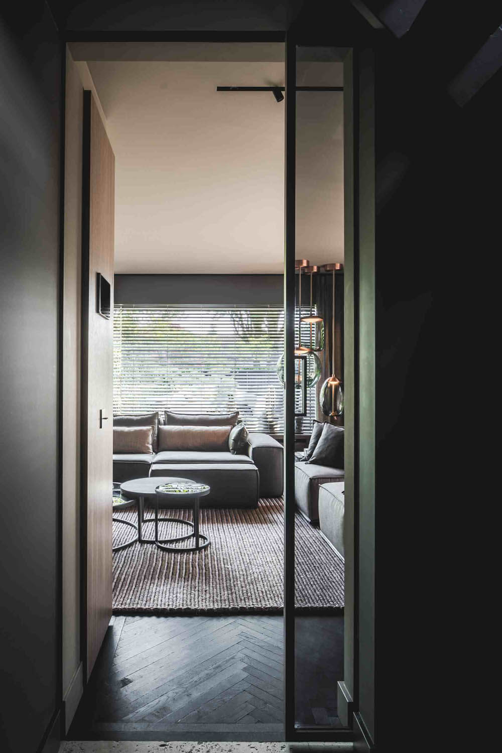 Framed view into a luxe living room with dark walls, L-shaped grey sofa, Brokis Knot pendants, and sunlit modern details.