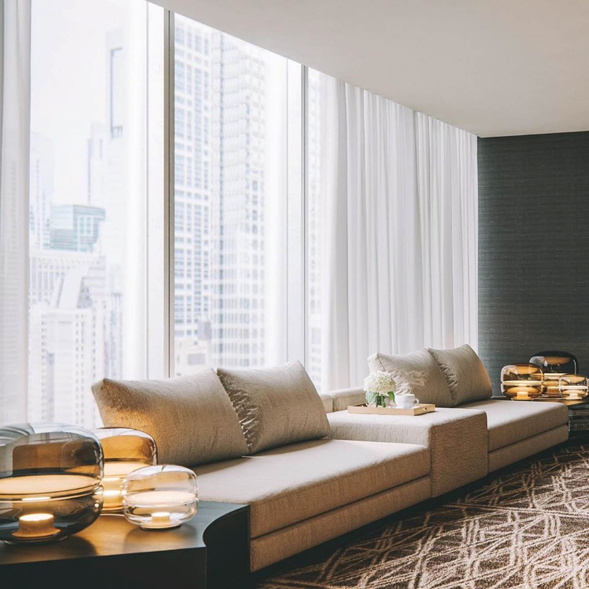A luxe lounge with two beige sofas, neutral cushions, and Brokis Macarons overlooks city views through sheer-draped windows.