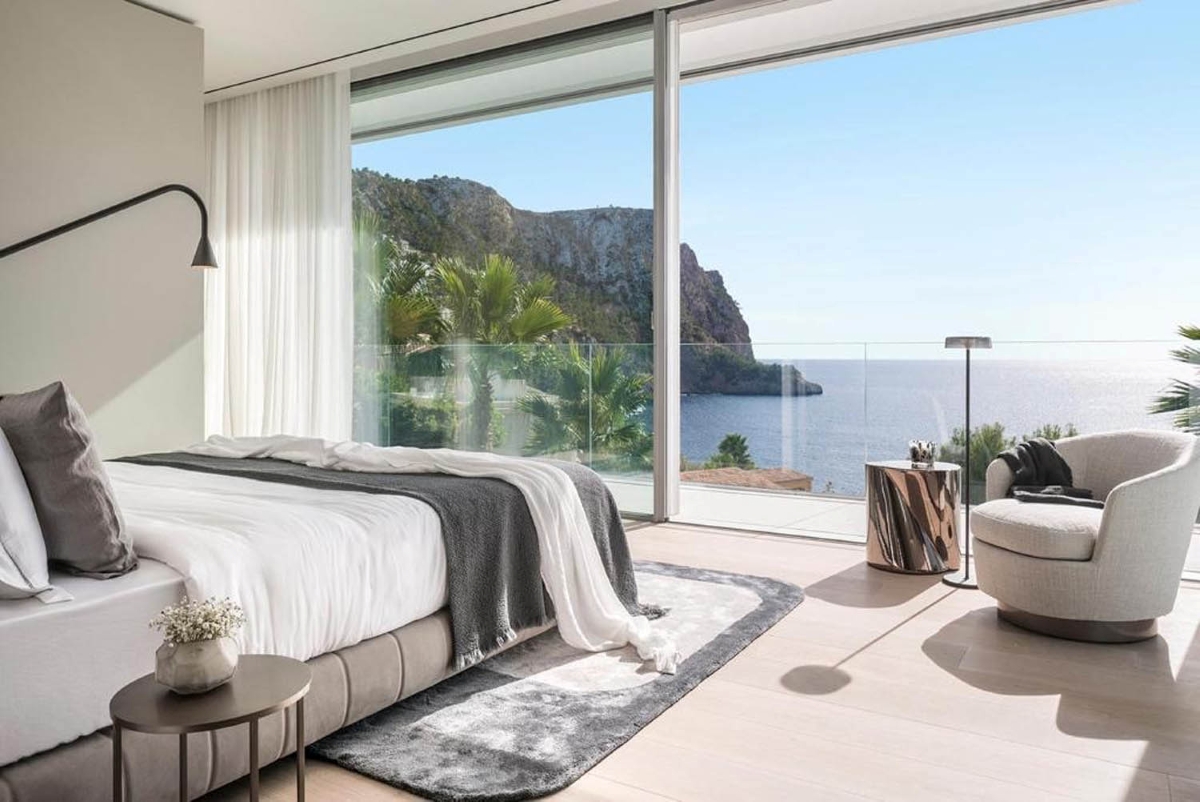 A sunlit, modern bedroom with a plush bed, luxury bedding, and ocean views through full-length windows.