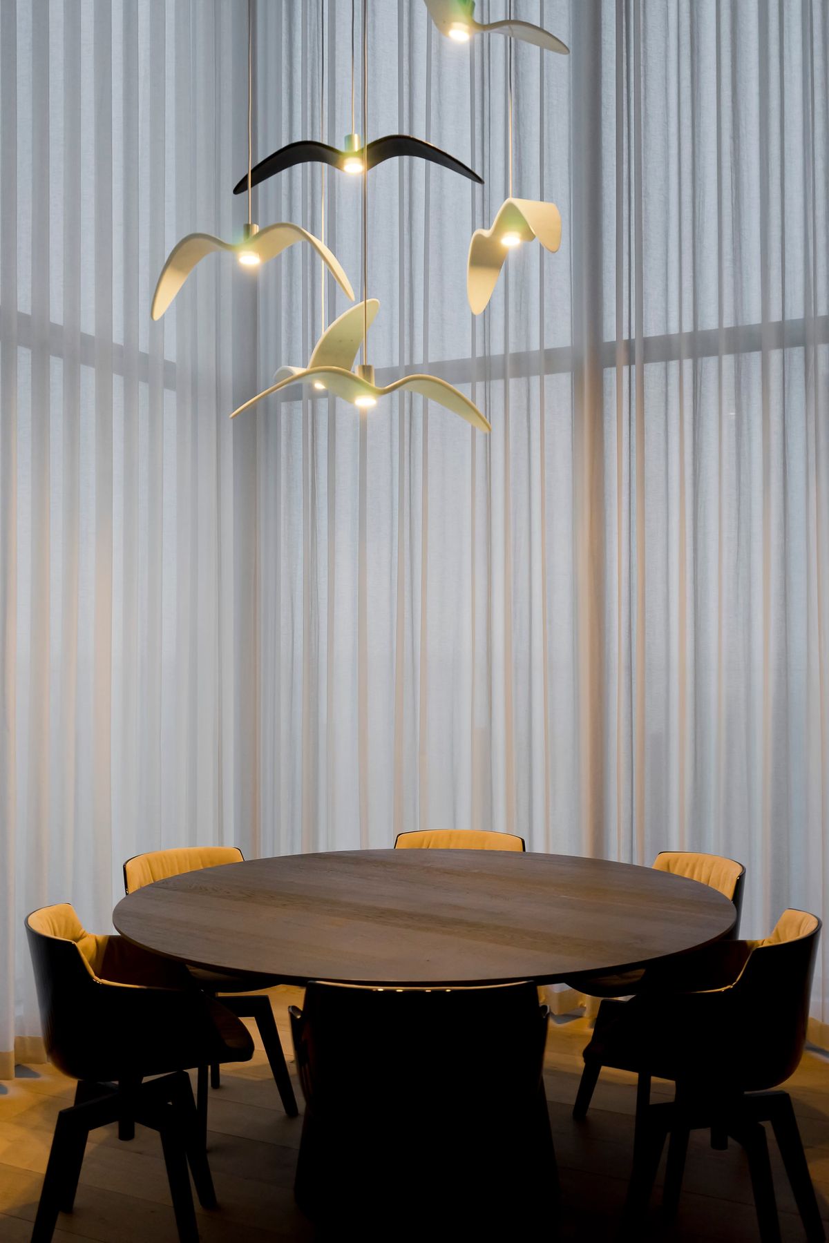 A round wooden table with six chairs sits beneath sculptural, Brokis Night Birds pendants in a room of sheer curtains.