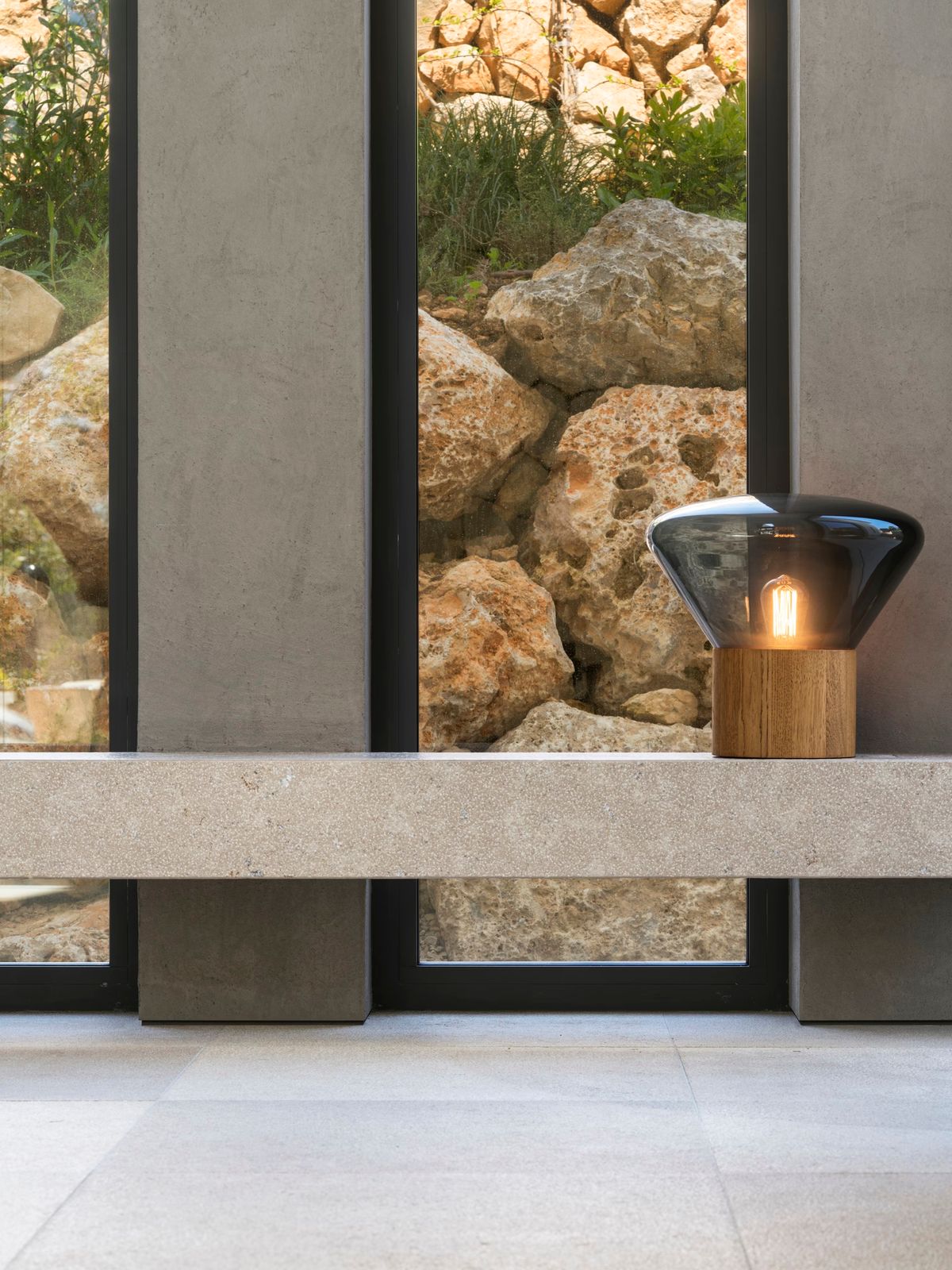 Brokis Muffins Wood 02 Table lamp with exposed bulb rests on stone before tall windows framing rocks and lush greenery.