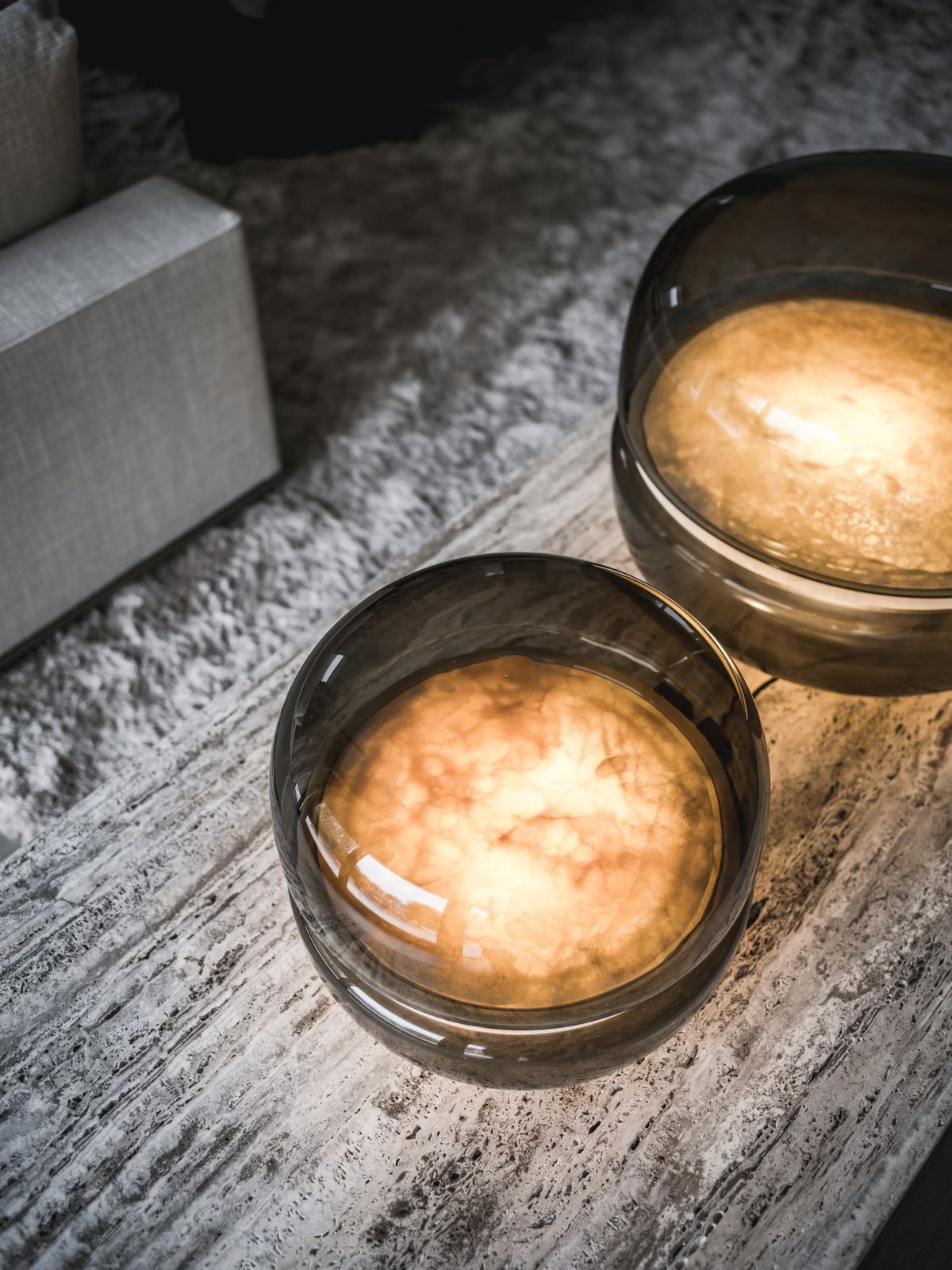 Two Brokis Macaron lamps in grey glass with soft onyx glow warmly on textured stone, exuding sculptural elegance.