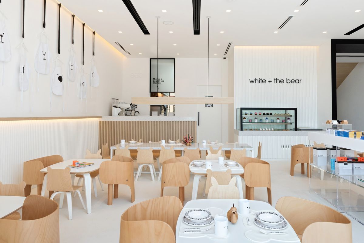 A minimalist children’s café with light wood chairs, and white tables, aprons on the wall.