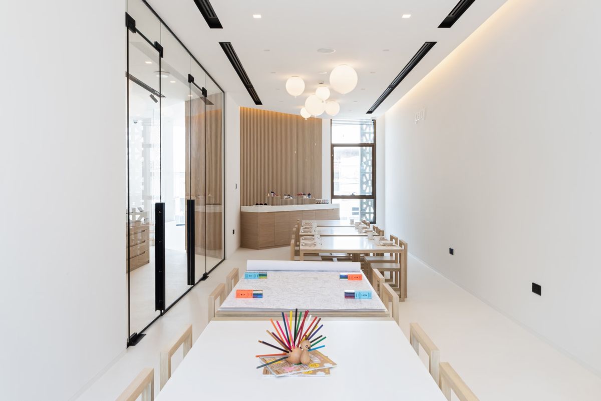 A luminous classroom with white walls and floors, light wood furniture, Brokis Memory ceiling lights, and a glass partition.