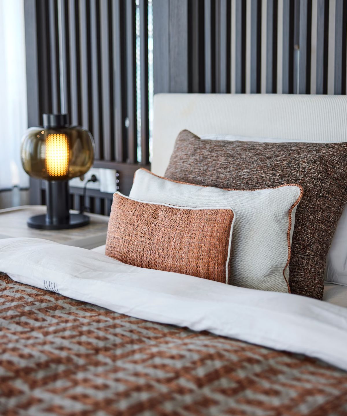 A bed with crisp white linen, brown and cream cushions, and a Brokis Bonbori Small Table Lamp with an amber crystal shade.