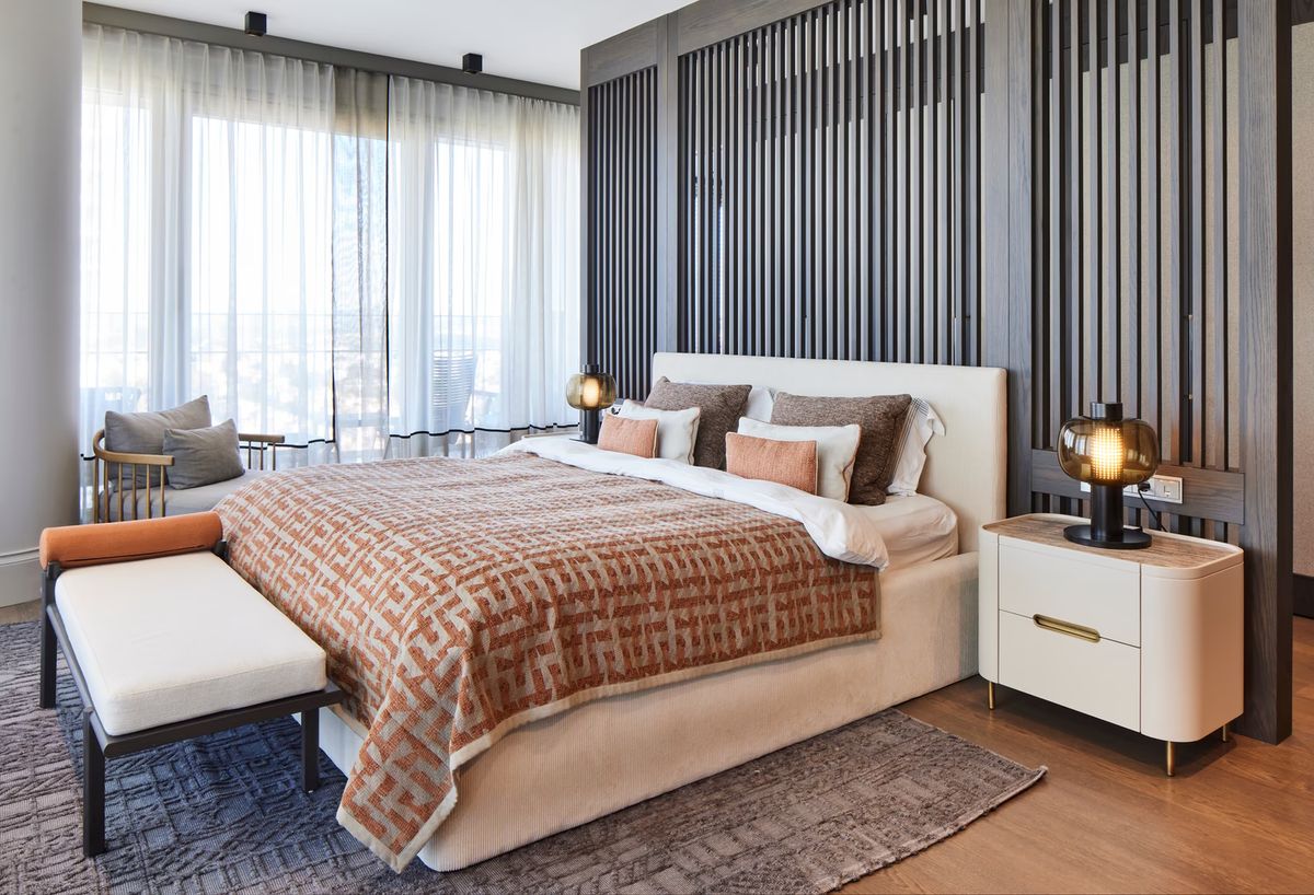 A luxury bedroom with a large bed in beige and orange, white bench, Brokis Bonbori Small, wood panelling, and sunlit windows.