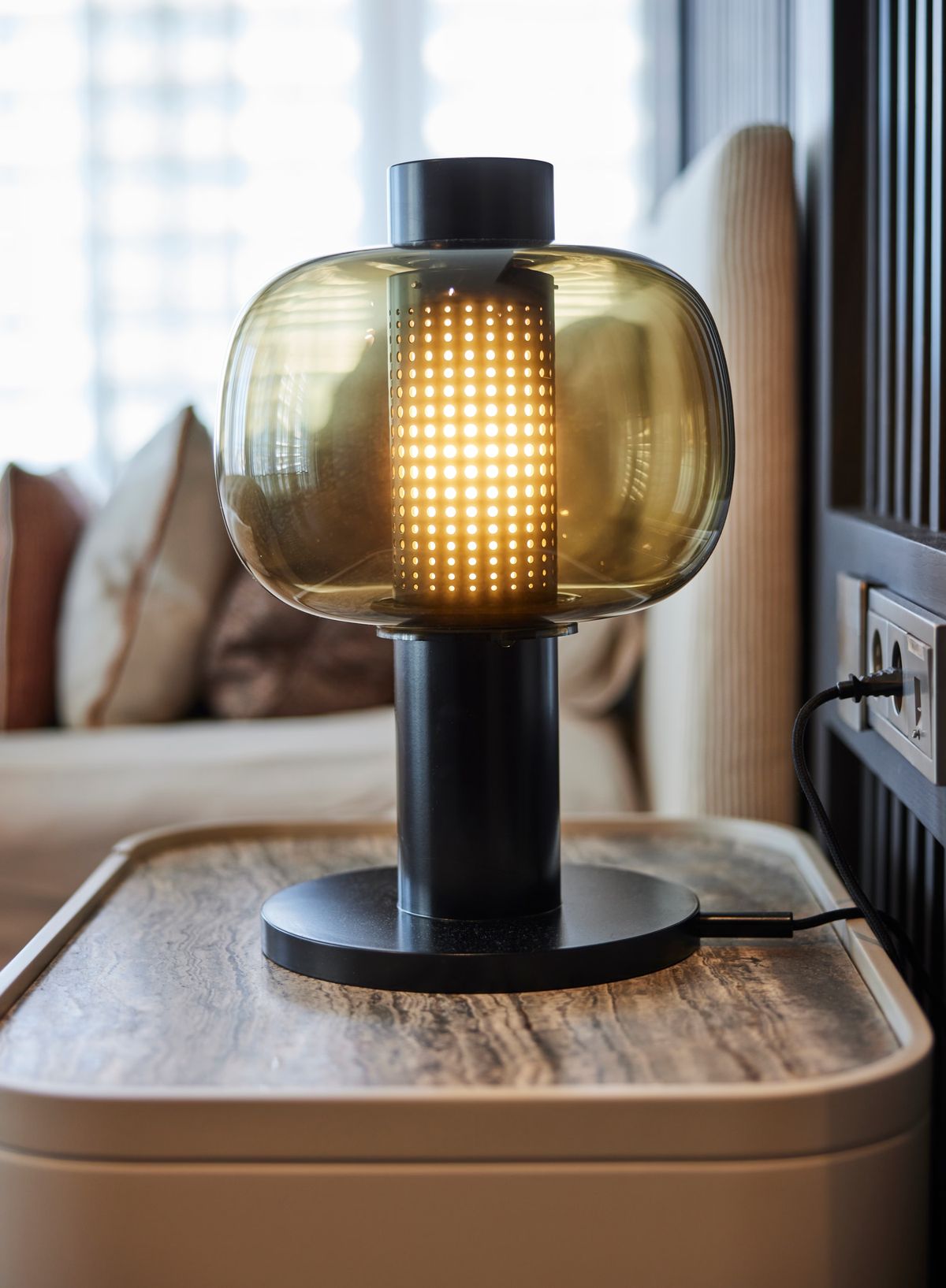 A Brokis Bonbori Small table lamp with a perforated shade glows on a marble-topped side table beside a plush-cushioned sofa.