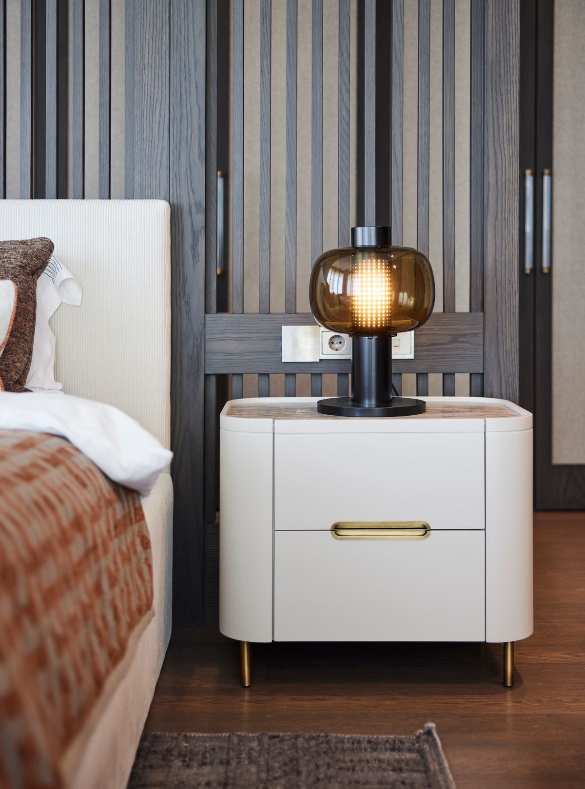 A luxe bedroom with white bedside table, gold accents, plush bedding, and a Brokis Bonbori Small glowing on wood panelling.