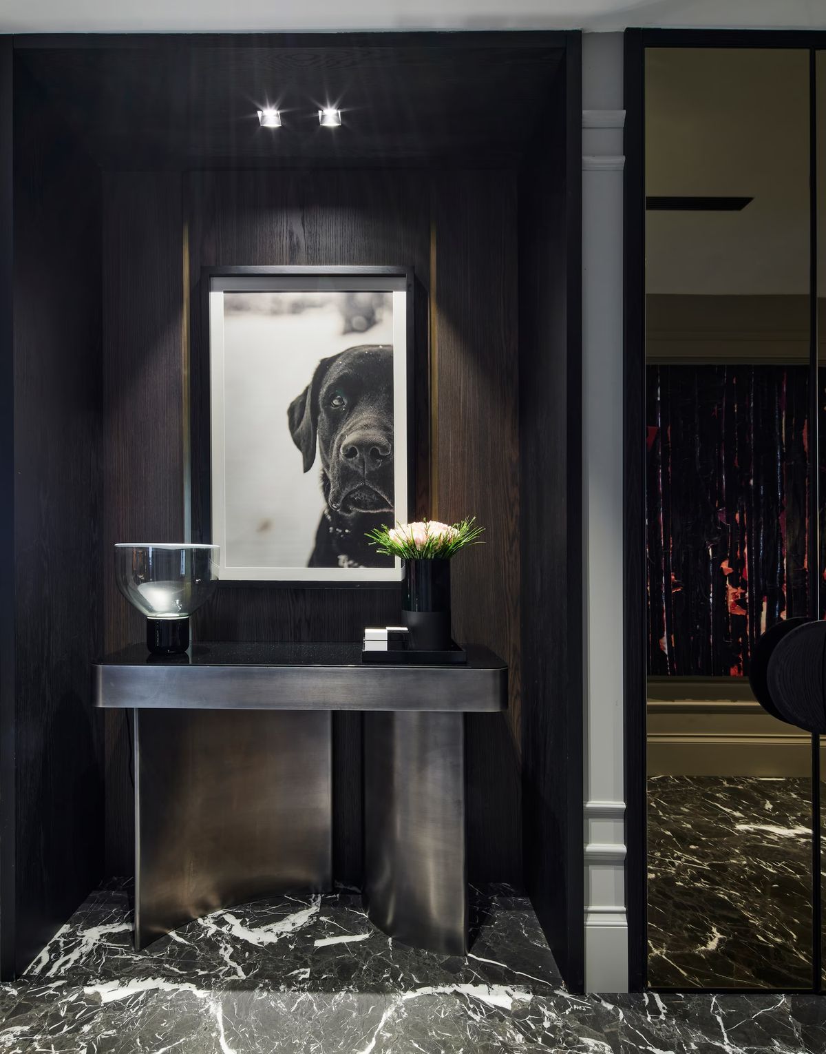 A black marble floor and dark alcove frame a metallic console with Brokis Lightline S, pale flowers, and illuminated art.