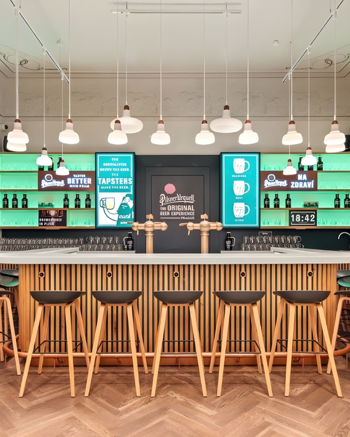 Pilsner Urquell Experience with a wooden counter, tall stools, Brokis Shadows pendants, and softly lit glass shelves.