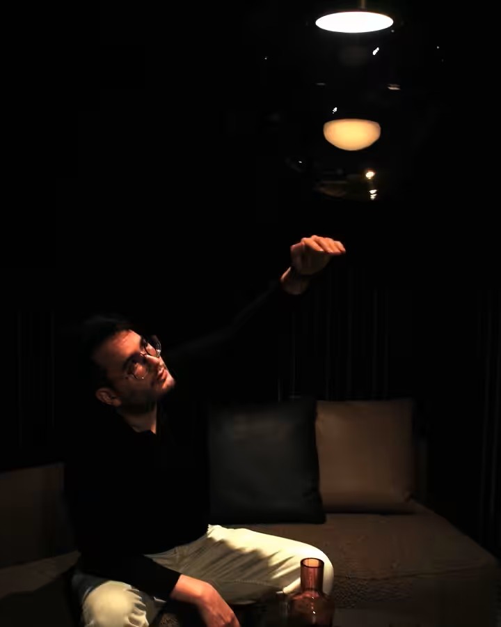 James Apichart Jarvis sits on a dim sofa, reaching for a Brokis Orbis pendant light that illuminates him softly.