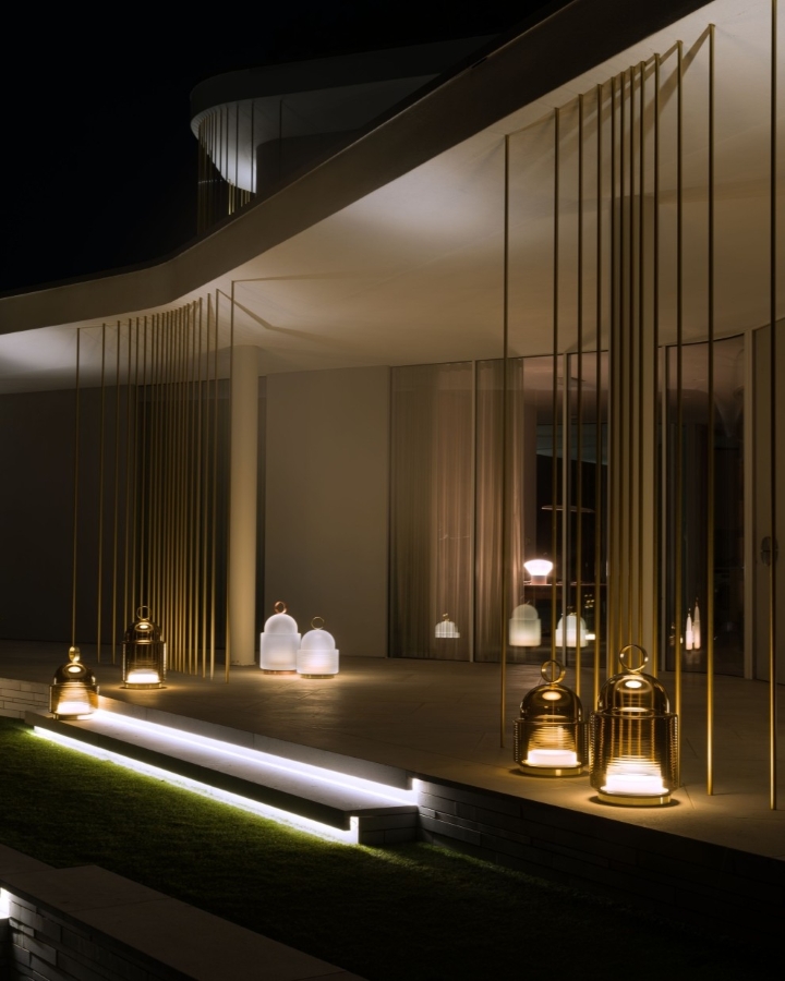 A modern building at night with gold vertical rods, illuminated lanterns, and soft white lights on the steps, creating an elegant, ambient atmosphere.