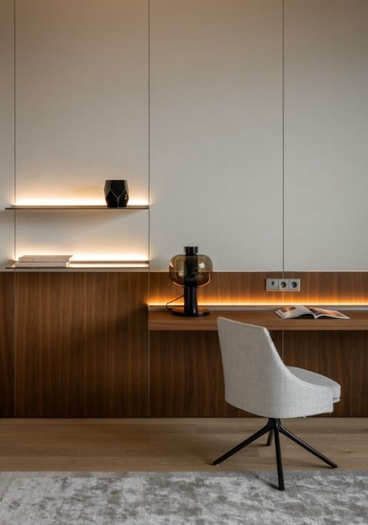 A modern minimalist workspace with a wooden desk, shelves, a grey swivel chair, a black table lamp, a black vase, and subtle built-in lighting against a light-coloured wall.