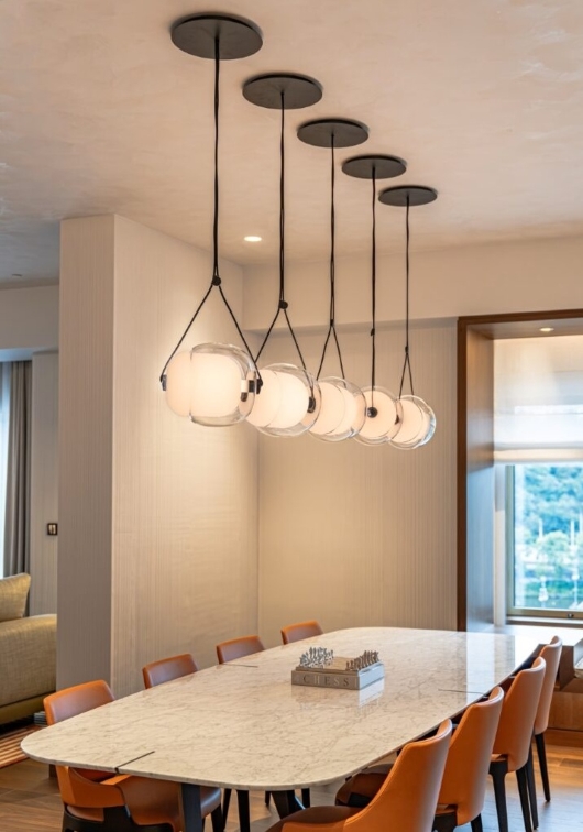 A modern dining room with a marble table, surrounded by orange chairs, and five spherical pendant lights hanging in a row above the table. A window with a view is visible in the background.
