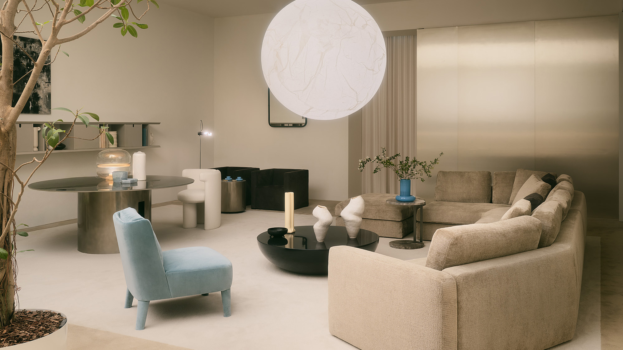 A modern living room with beige corner sofa, a light blue armchair, round black coffee table, dining area with a dark table and white chairs, large round pendant light, neutral walls, and minimalist decor.