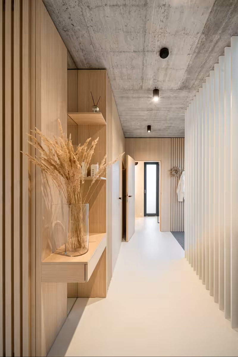 A modern hallway with light wood panelling, a concrete ceiling, white flooring, built-in shelves holding a glass vase with dried grasses, and a door at the end letting in natural light.