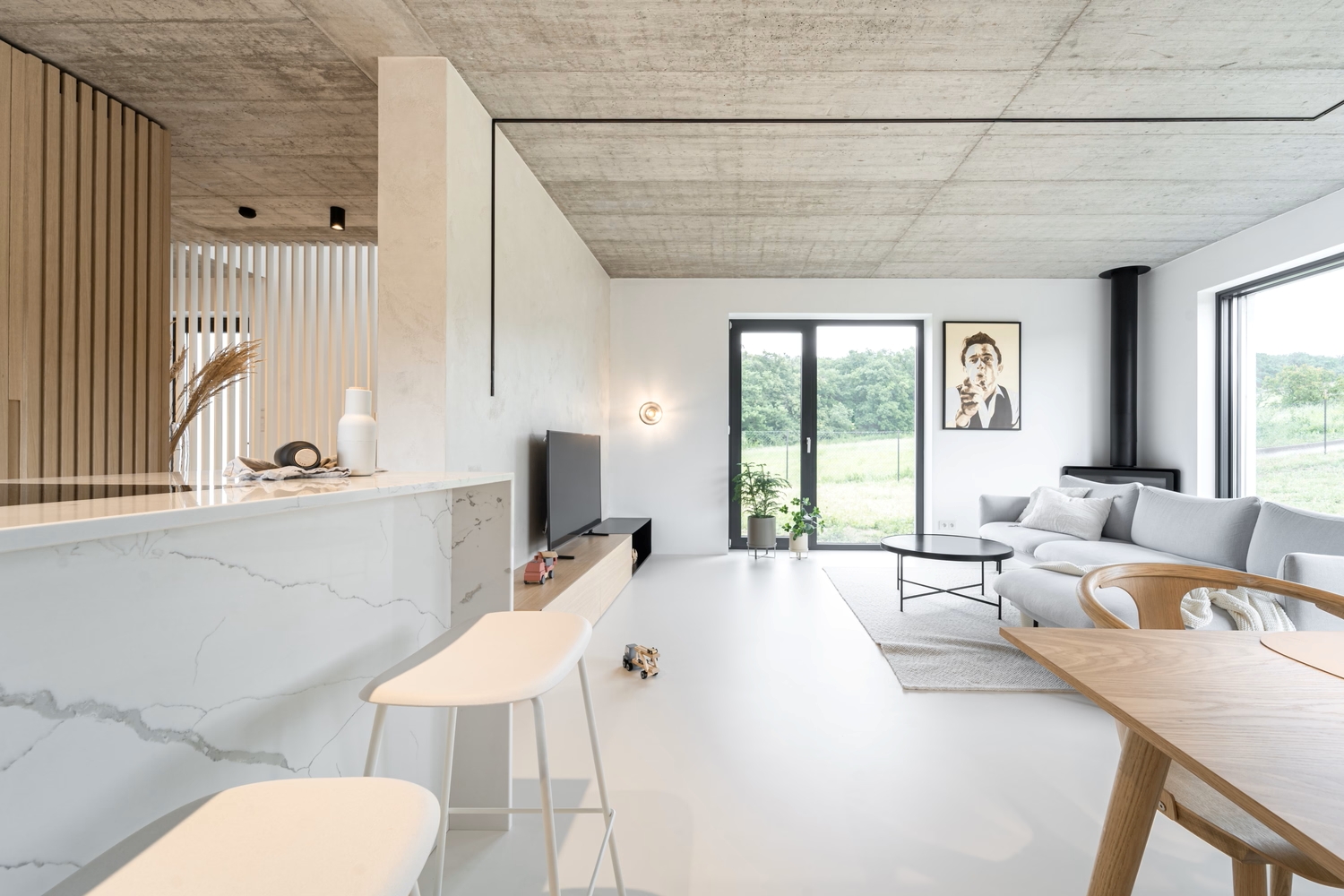 Modern minimalist living room with large windows, a grey corner sofa, wall art, and a TV. Light wood and white furniture, marble worktop, and green views outside create a bright, airy atmosphere.