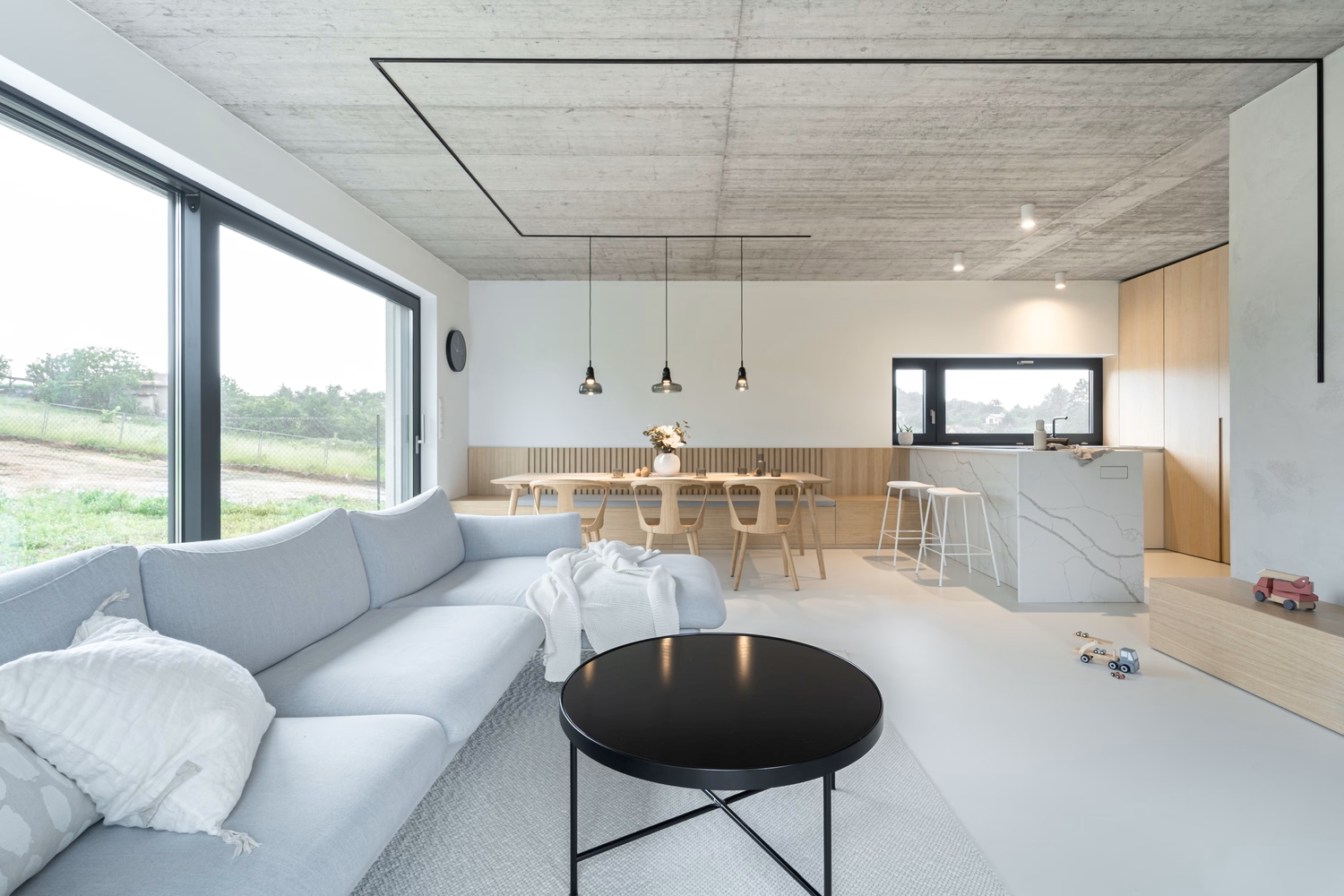 Modern open-plan living area with large windows, light grey corner sofa, round black coffee table, dining table with wooden chairs, minimalist kitchen, and toys scattered on the floor. Outdoor greenery visible through windows.
